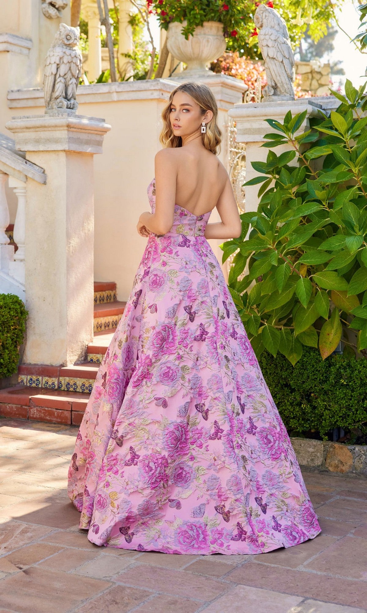 A woman wearing the Juliet JT2735H long prom dress stands outdoors by stairs and greenery, looking over her shoulder at the camera.